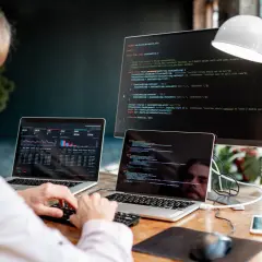 Mężczyzna przy biurku z dwoma laptopami i monitorem wyświetlającymi kod.