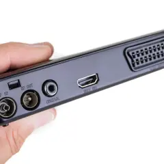 Dłoń trzyma czarny dekoder z portami RF IN, RF OUT, COAXIAL, HDMI OUT i SCART.