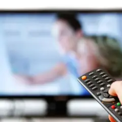 Dłoń trzymająca pilot do telewizora przed ekranem.