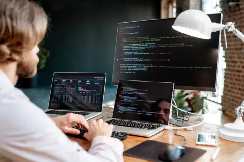 Mężczyzna przy biurku z dwoma laptopami i monitorem wyświetlającymi kod.