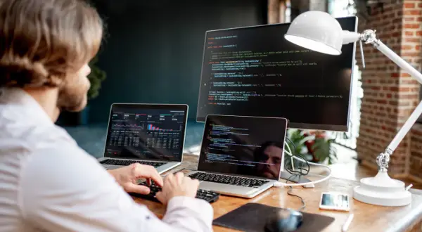 Mężczyzna przy biurku z dwoma laptopami i monitorem wyświetlającymi kod.