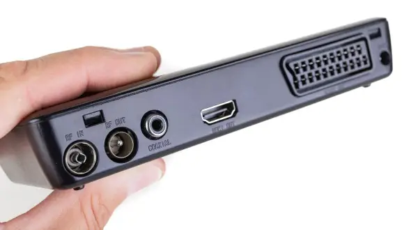Dłoń trzyma czarny dekoder z portami RF IN, RF OUT, COAXIAL, HDMI OUT i SCART.