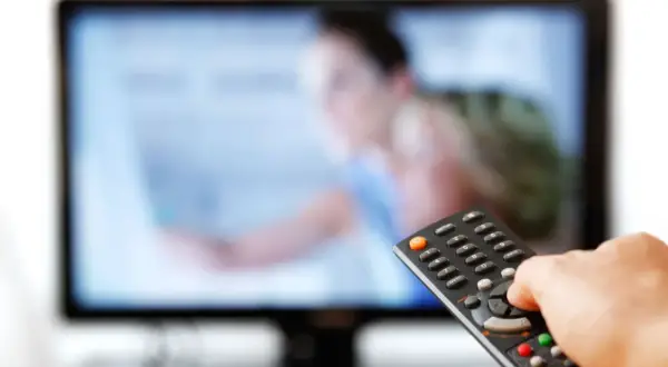 Dłoń trzymająca pilot do telewizora przed ekranem.