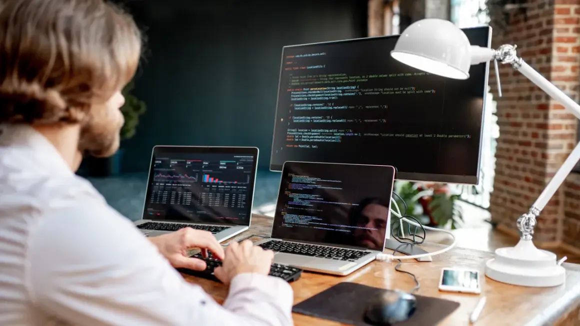 Mężczyzna przy biurku z dwoma laptopami i monitorem wyświetlającymi kod.
